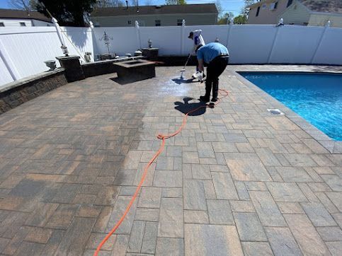 Person pressure washes a brick patio beside a blue swimming pool.