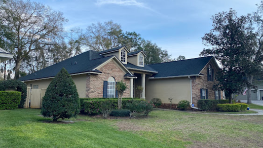Suburban single-story brick house with beige siding, dark roof, and manicured lawn under a blue sky