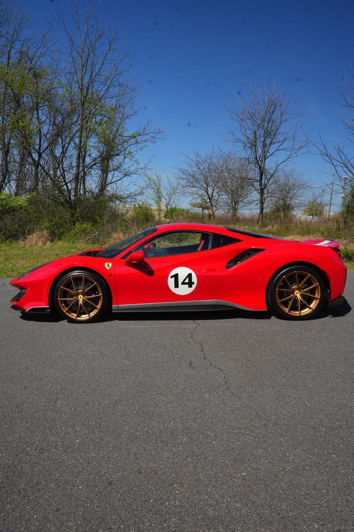 Red sports car with gold wheels and number 14 parked on a road beside trees under a blue sky