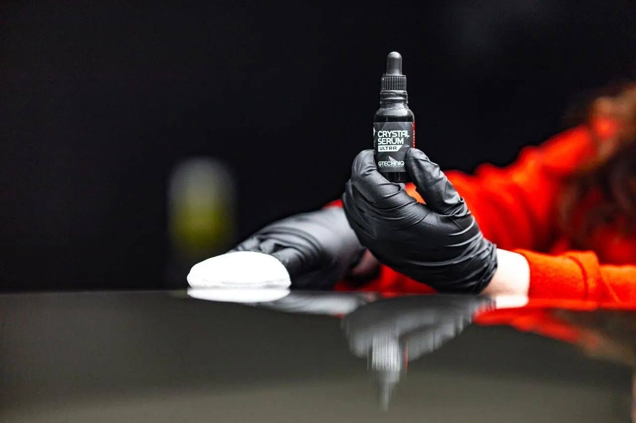 Person in black gloves holding a bottle of detailing product, applying it to a car surface with a white applicator.