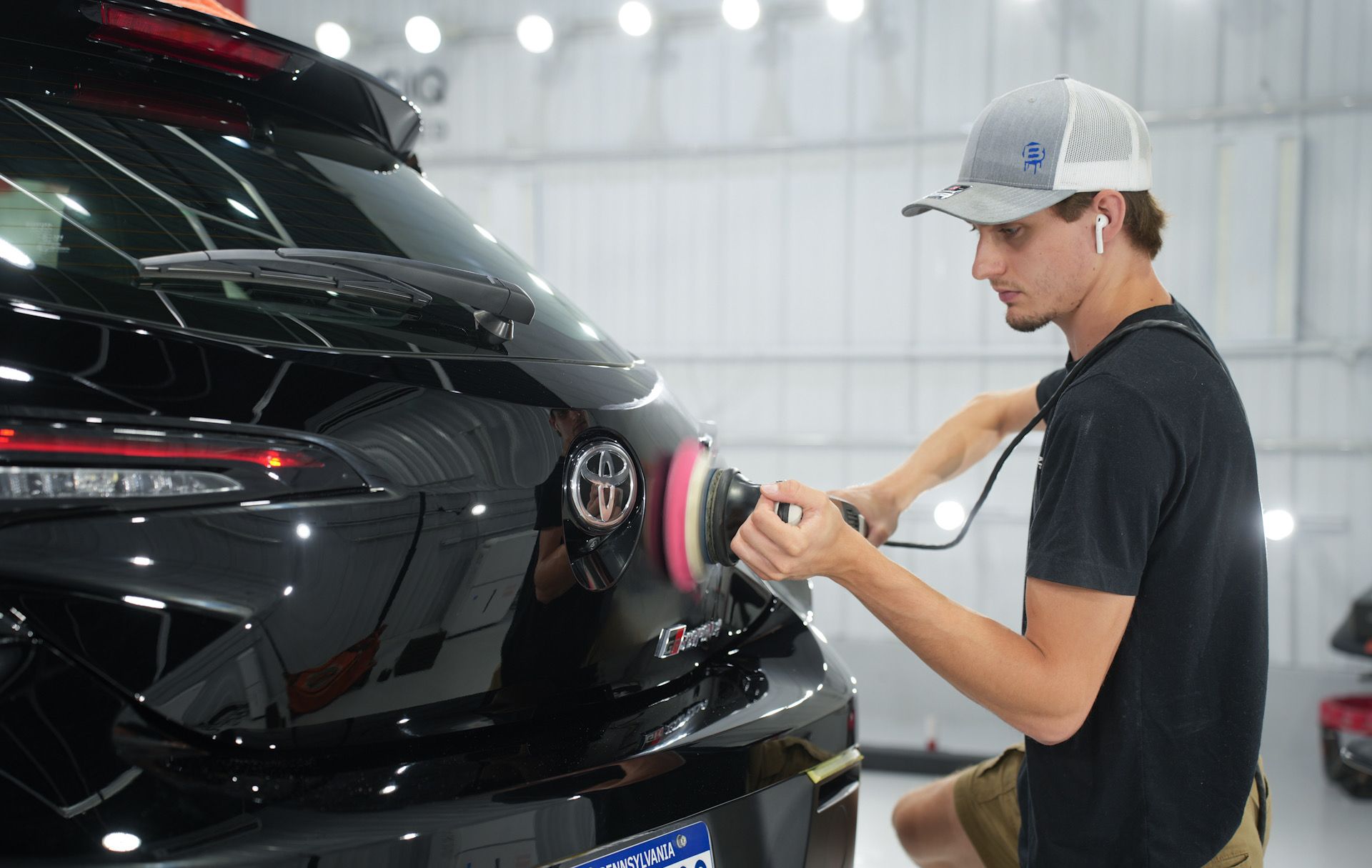 Man polishing the black rear end of a car in a well-lit shop, wearing a hat and headphones.