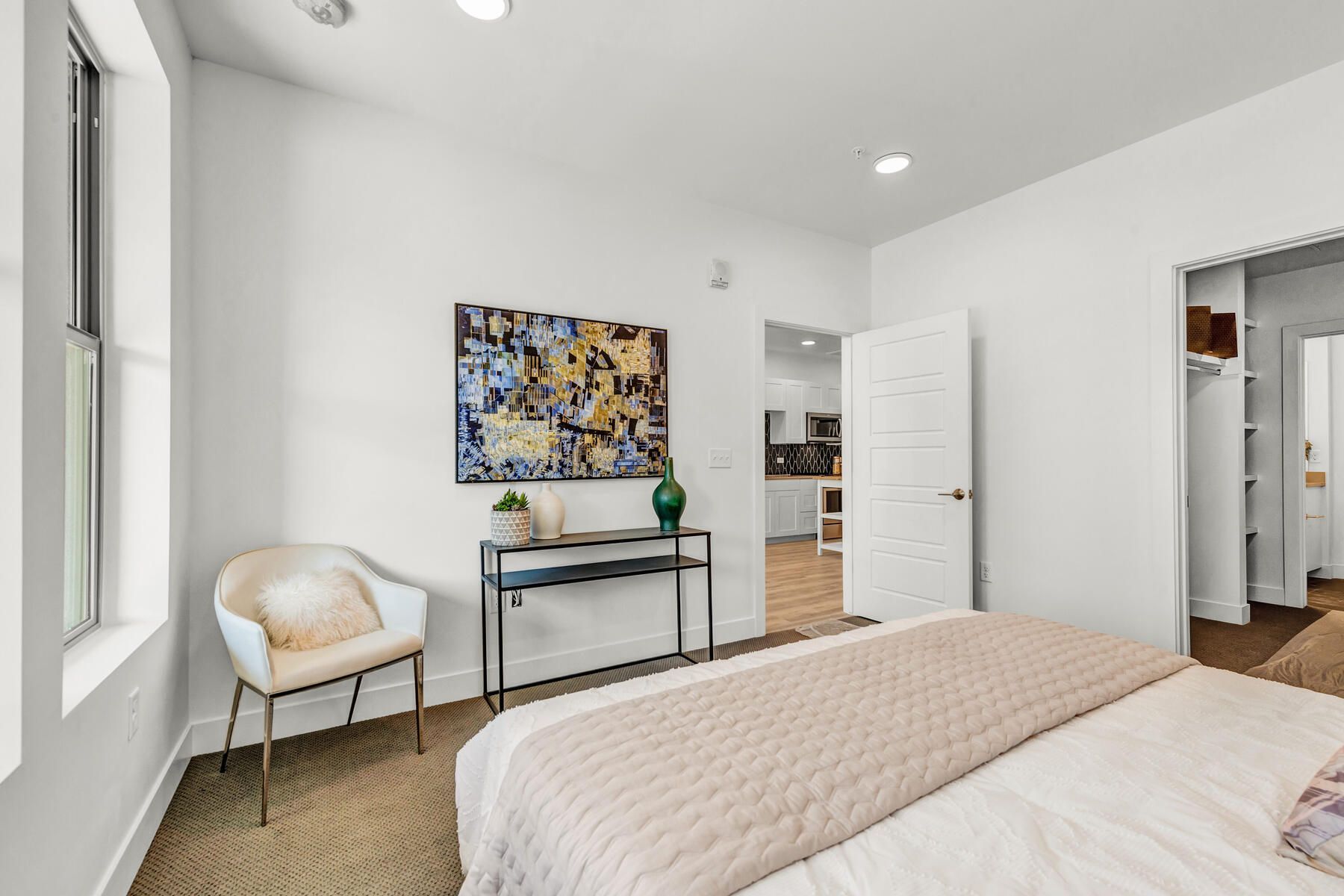 Bedroom with white walls, art, a chair, and a bed with a neutral-colored blanket. Door to closet on right.