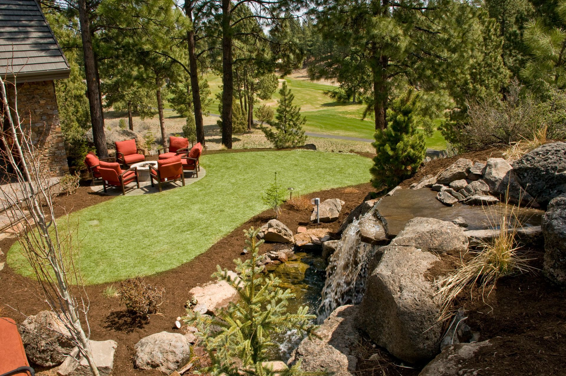 A backyard landscape with a small waterfall, green lawn, red chairs around a fire pit, and trees.