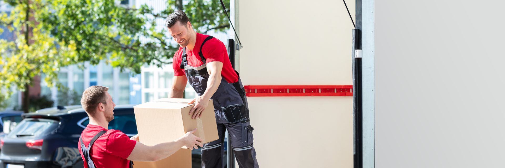 Two movers in red shirts loading a cardboard box into a moving truck outdoors. Two movers in red shirts loading a cardboard box into a moving truck outdoors.