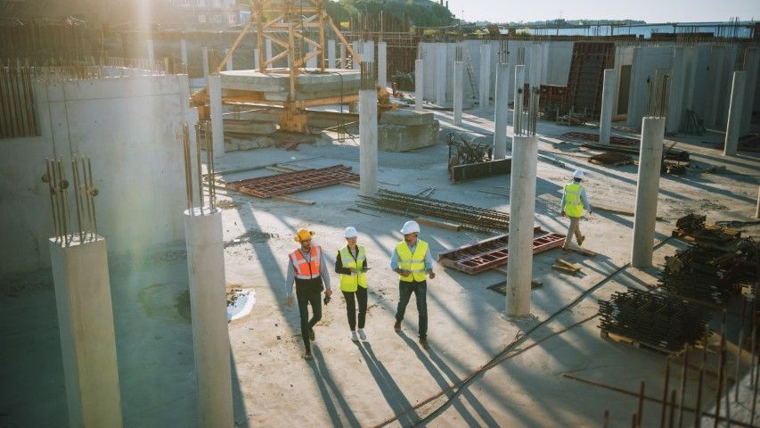Three people in construction gear walk through an unfinished building site with concrete pillars and sunlight overhead.