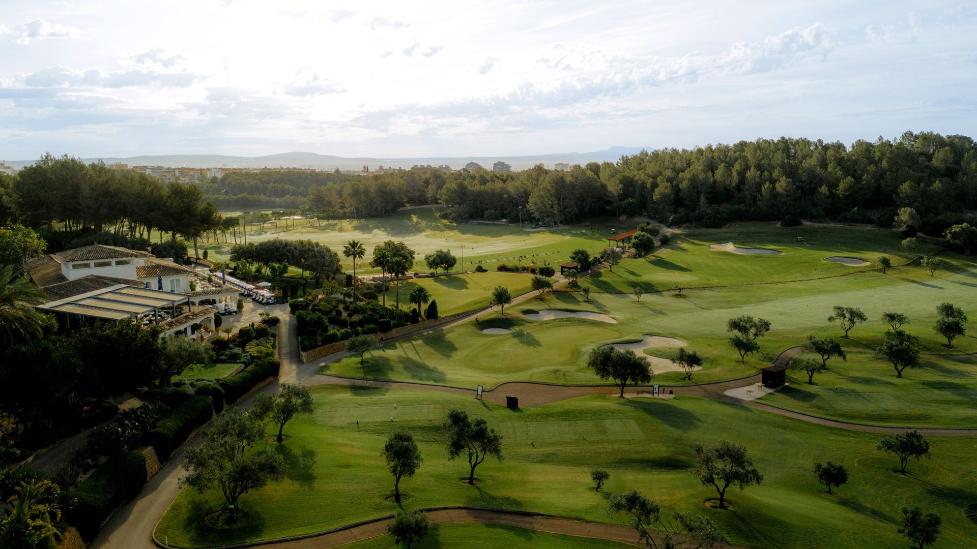 Aerial view of a scenic golf resort, Arabella Golf, with lush green fairways, sand bunkers, and surrounding trees under a hazy sky.