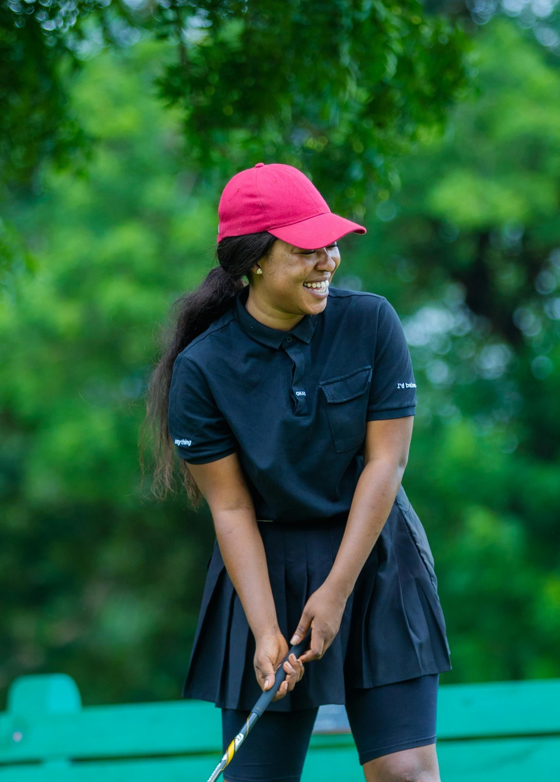 Woman in black golf attire and red cap smiles while swinging a golf club outdoors.