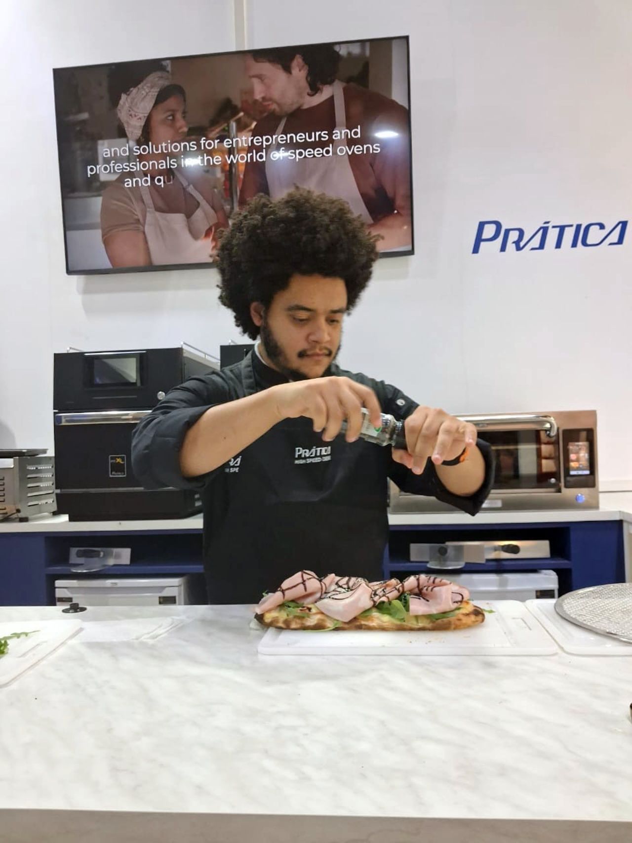 Chef adding seasoning to a sandwich on a counter with appliances and a TV in the background.