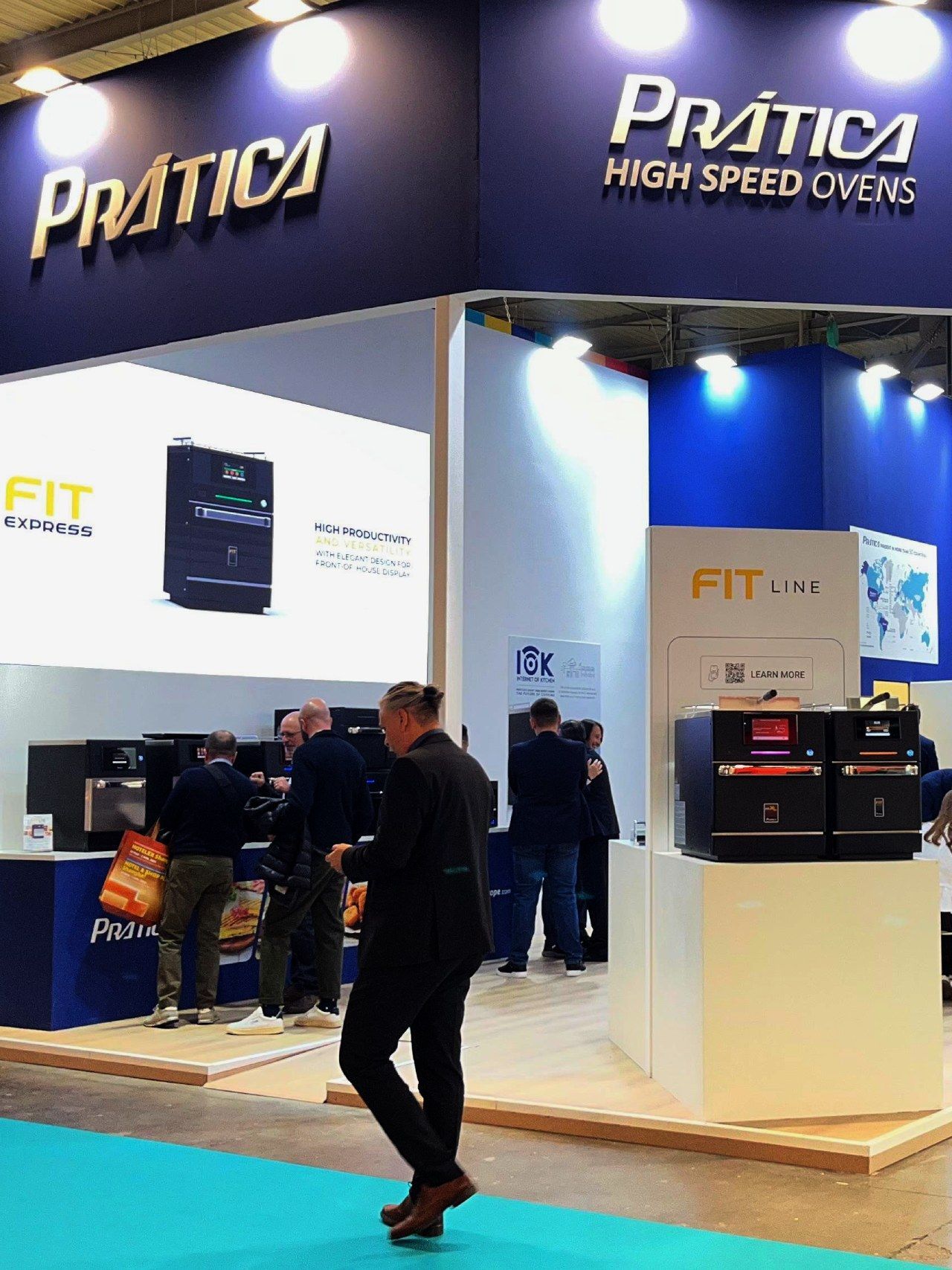 Prática High Speed Ovens booth with people, ovens, and a man walking in a blue and white setting.