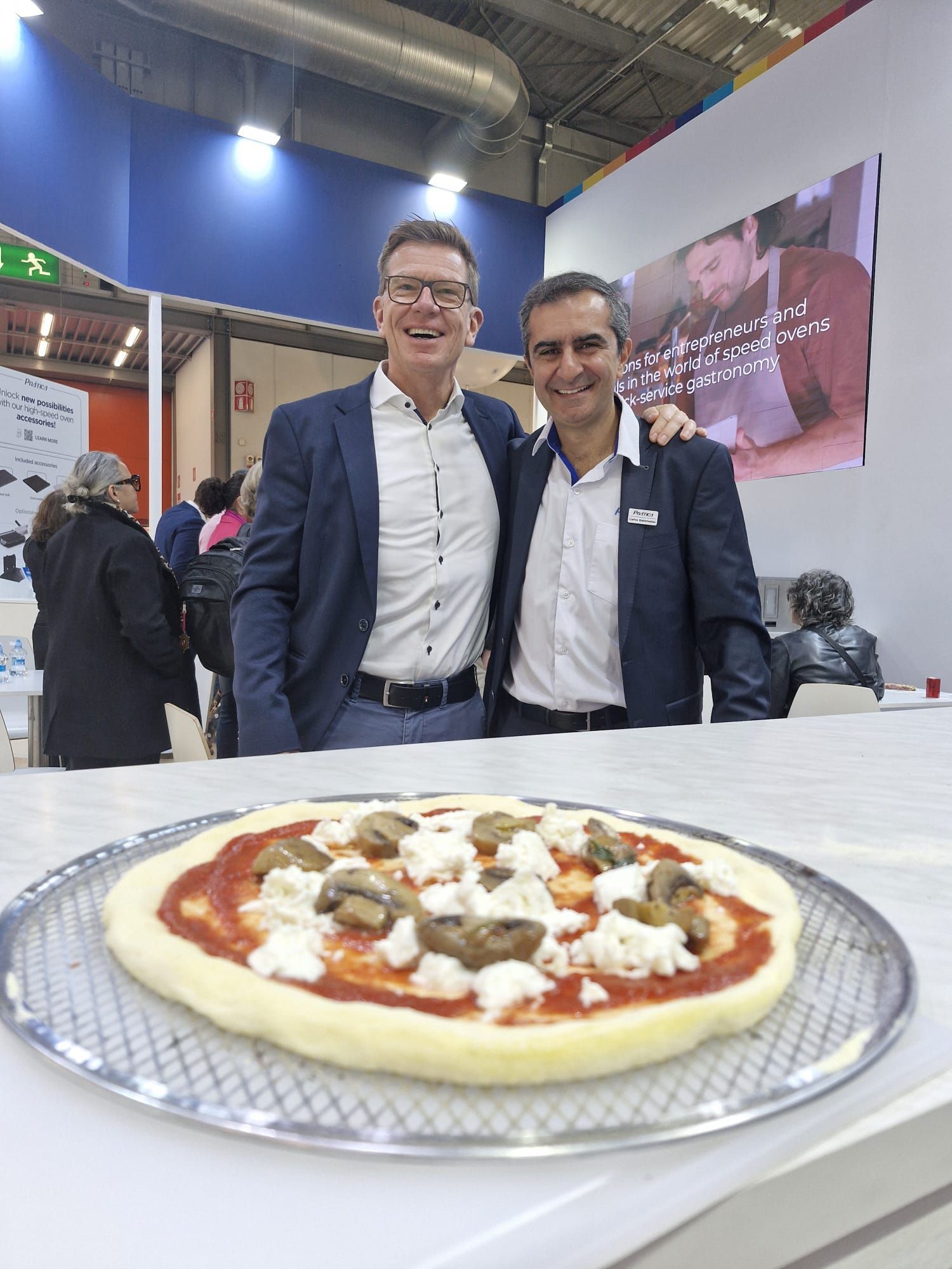 Two men pose with a pizza, at a trade show.