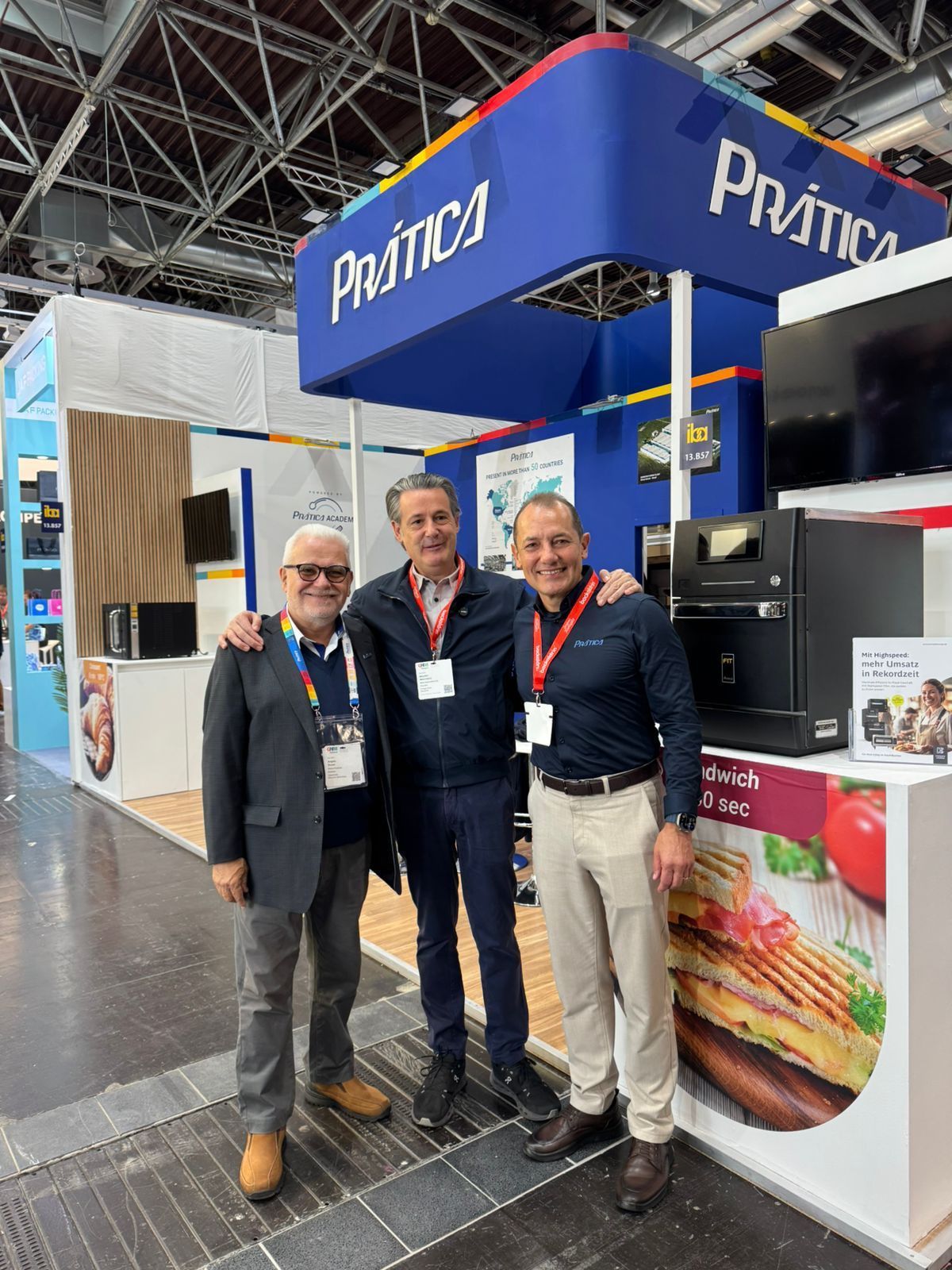 Three men pose at a trade show booth for Pratíca. One man is wearing a jacket, one a blue jacket and one is in a blue polo.