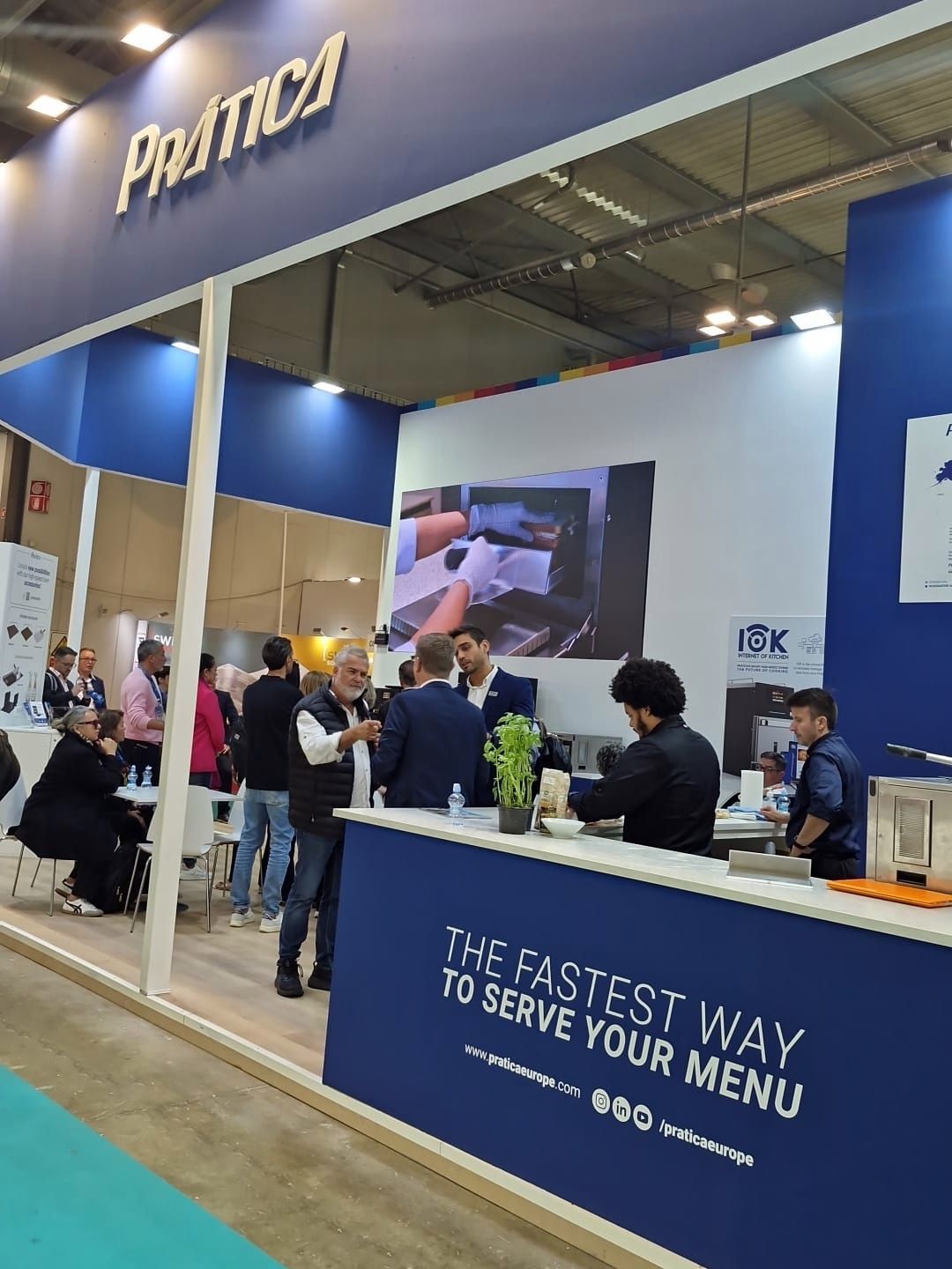Prática booth at a trade show; people interacting, blue and white branding, text 