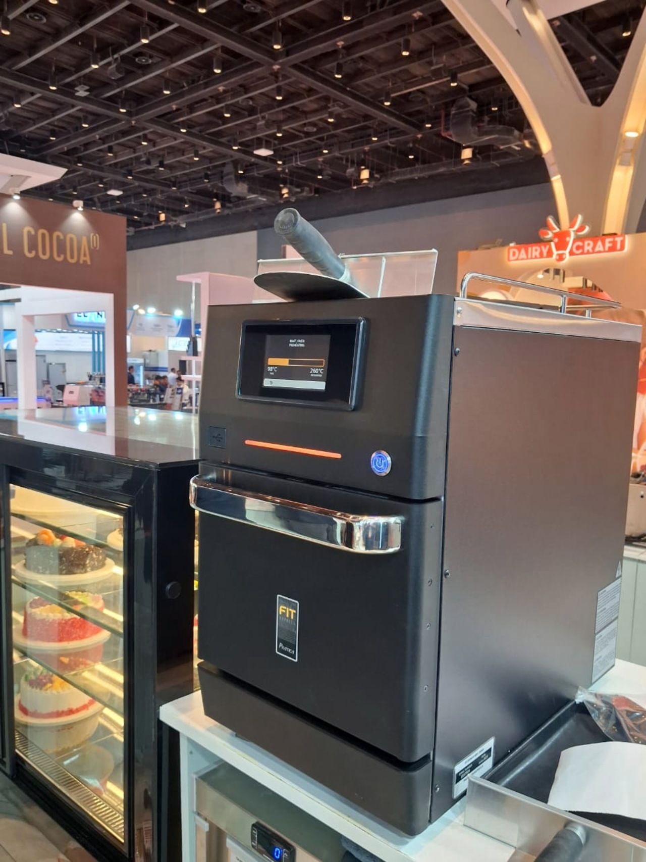 Black commercial kitchen appliance with a digital display, scoop, and display case of food at a trade show.