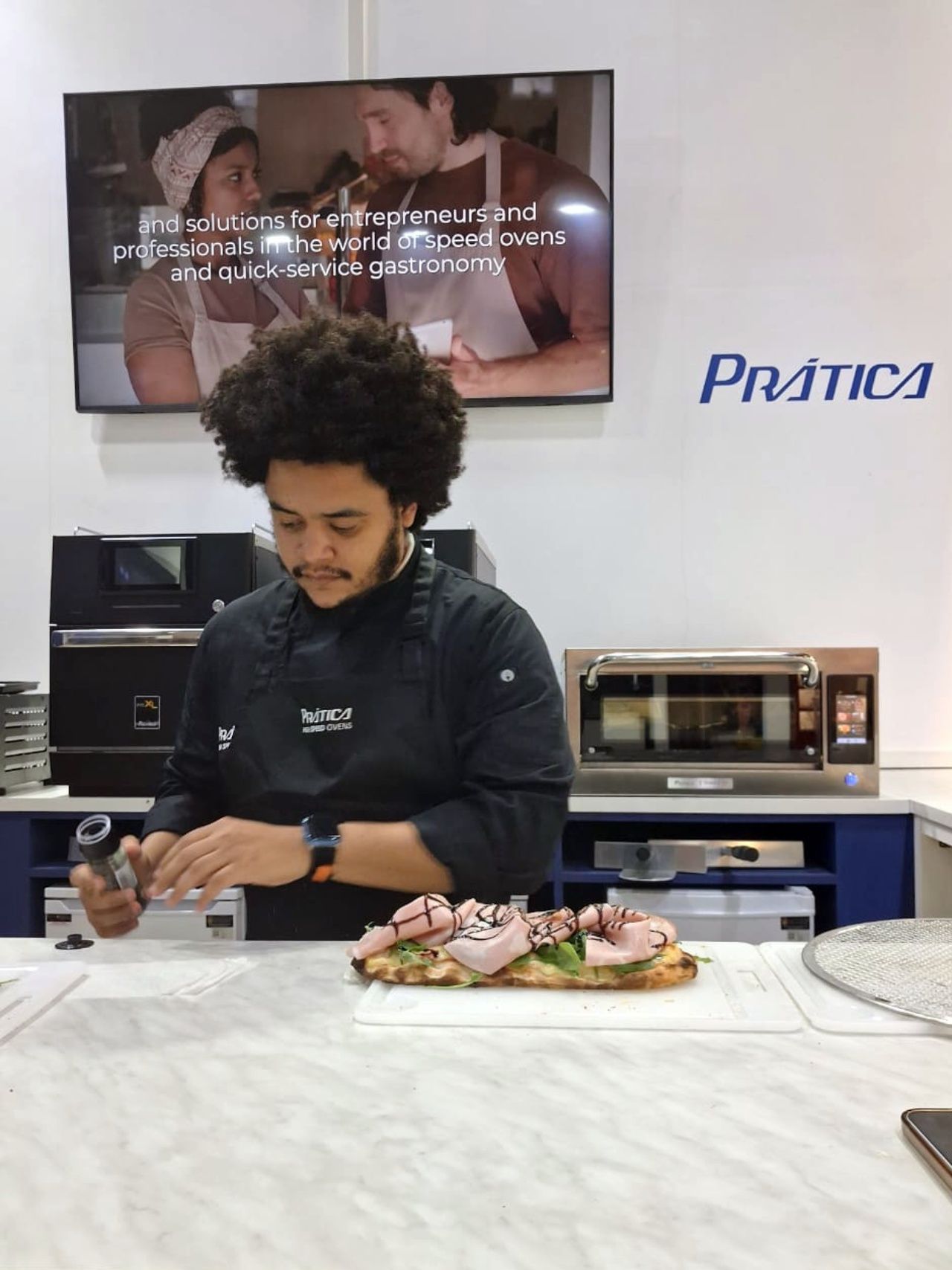 Chef preparing a sandwich at a Prática appliance display, with oven visible.