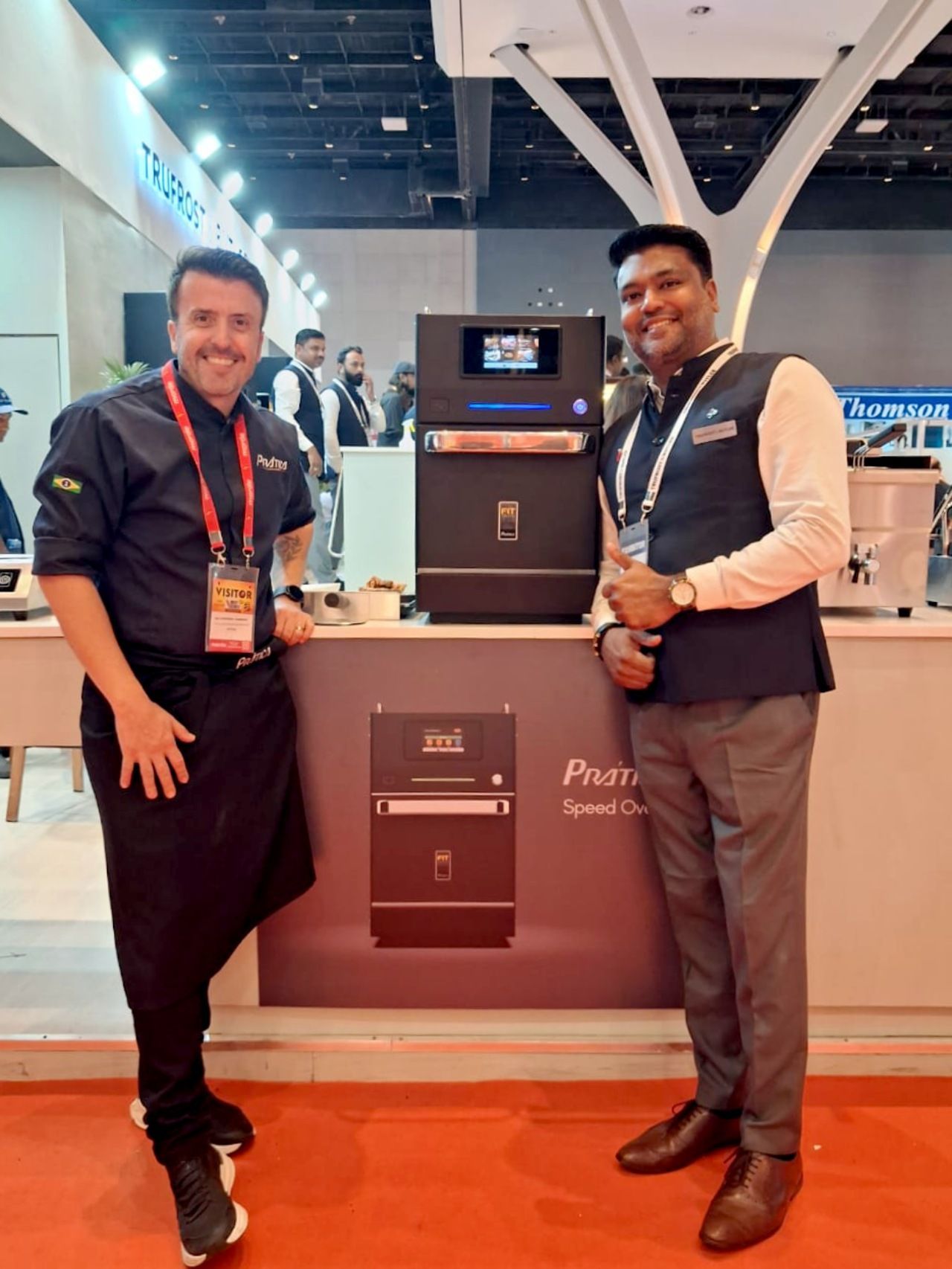 Two men pose by a black appliance at a trade show. One in chef attire, the other in a vest. Sign says 