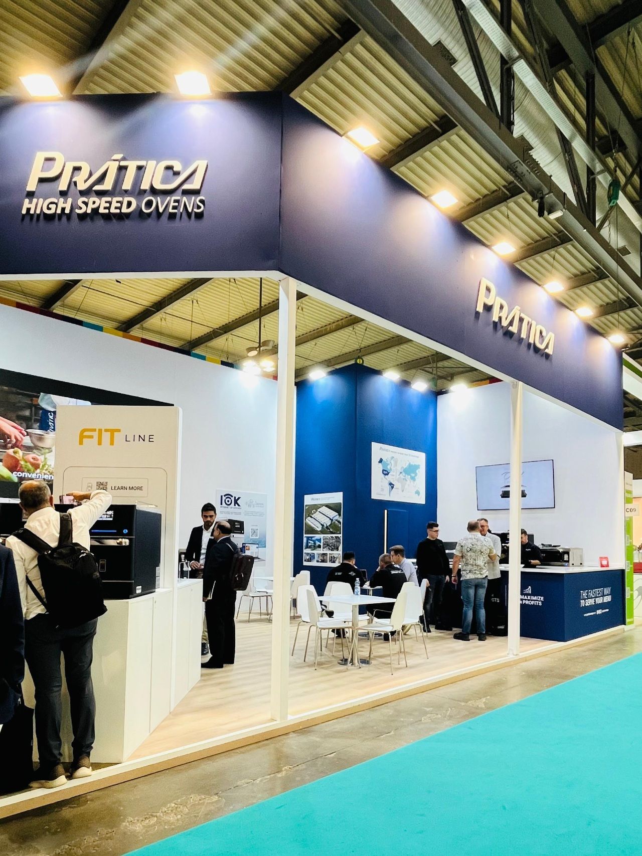 Prática High Speed Ovens booth at a trade show. Blue and white colors, people browsing, chairs and tables.