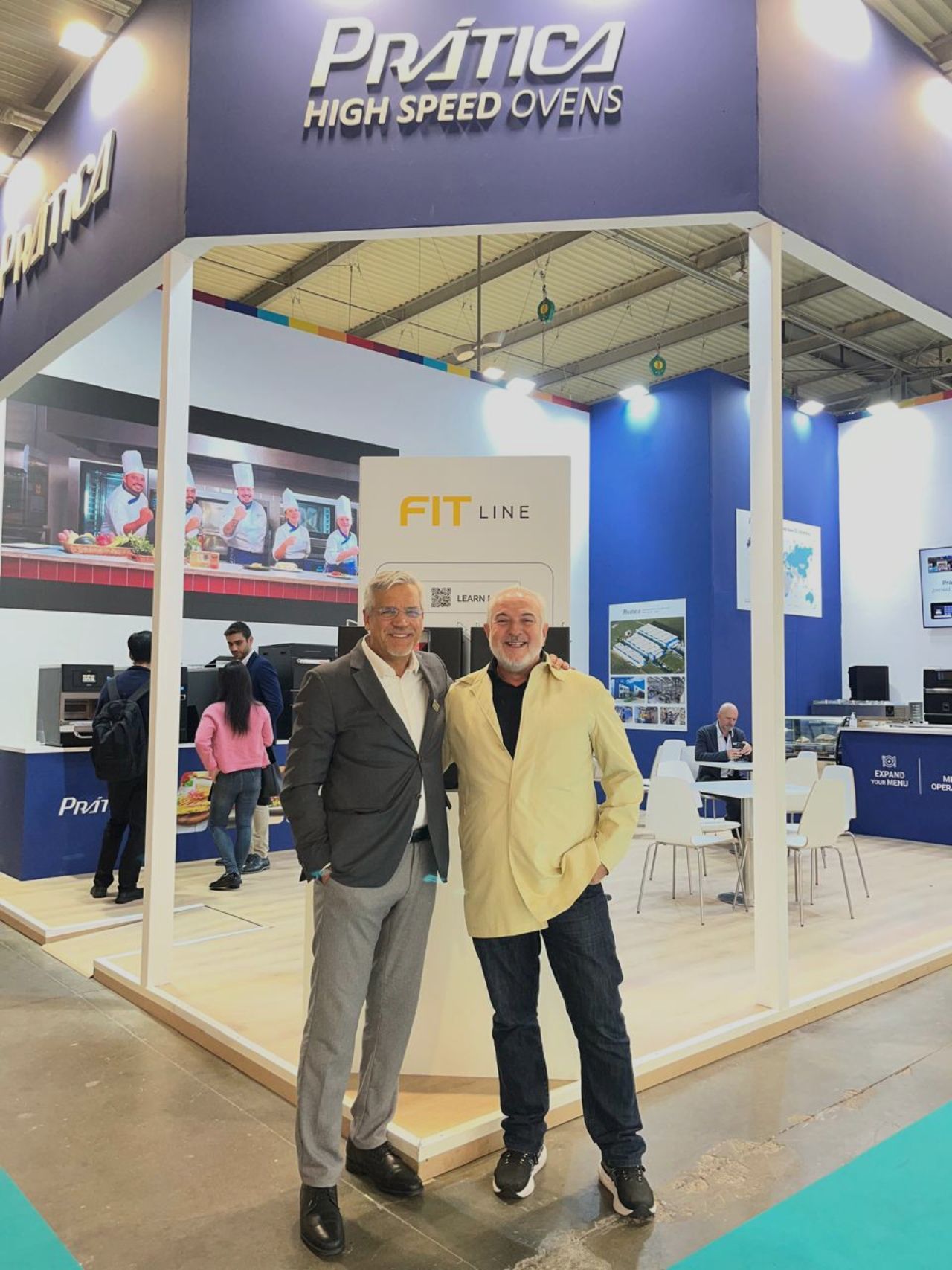 Two men pose in front of a trade show booth promoting Prática High Speed Ovens. The booth has blue and white colors.