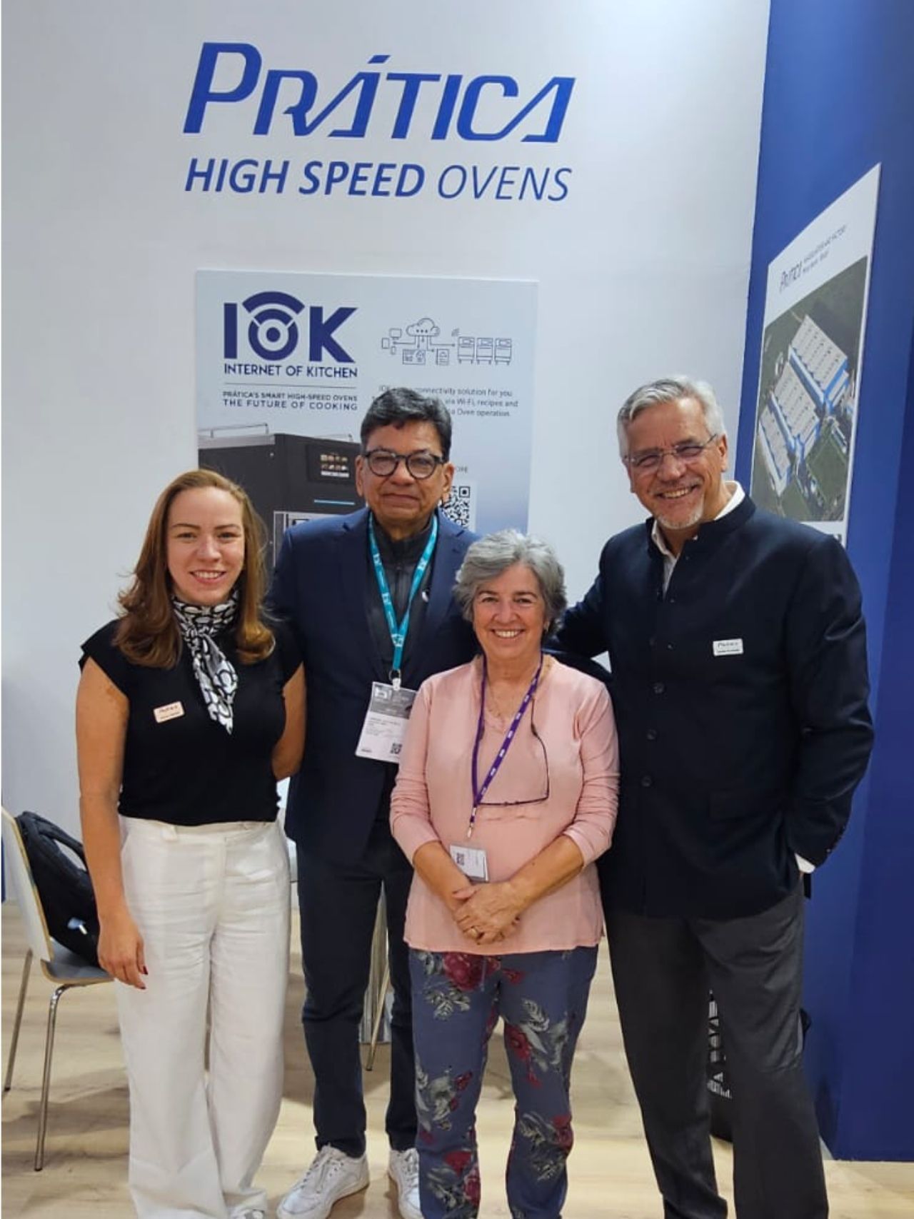Four people pose in front of a “Prática High Speed Ovens” booth. They are smiling. A poster is behind them.