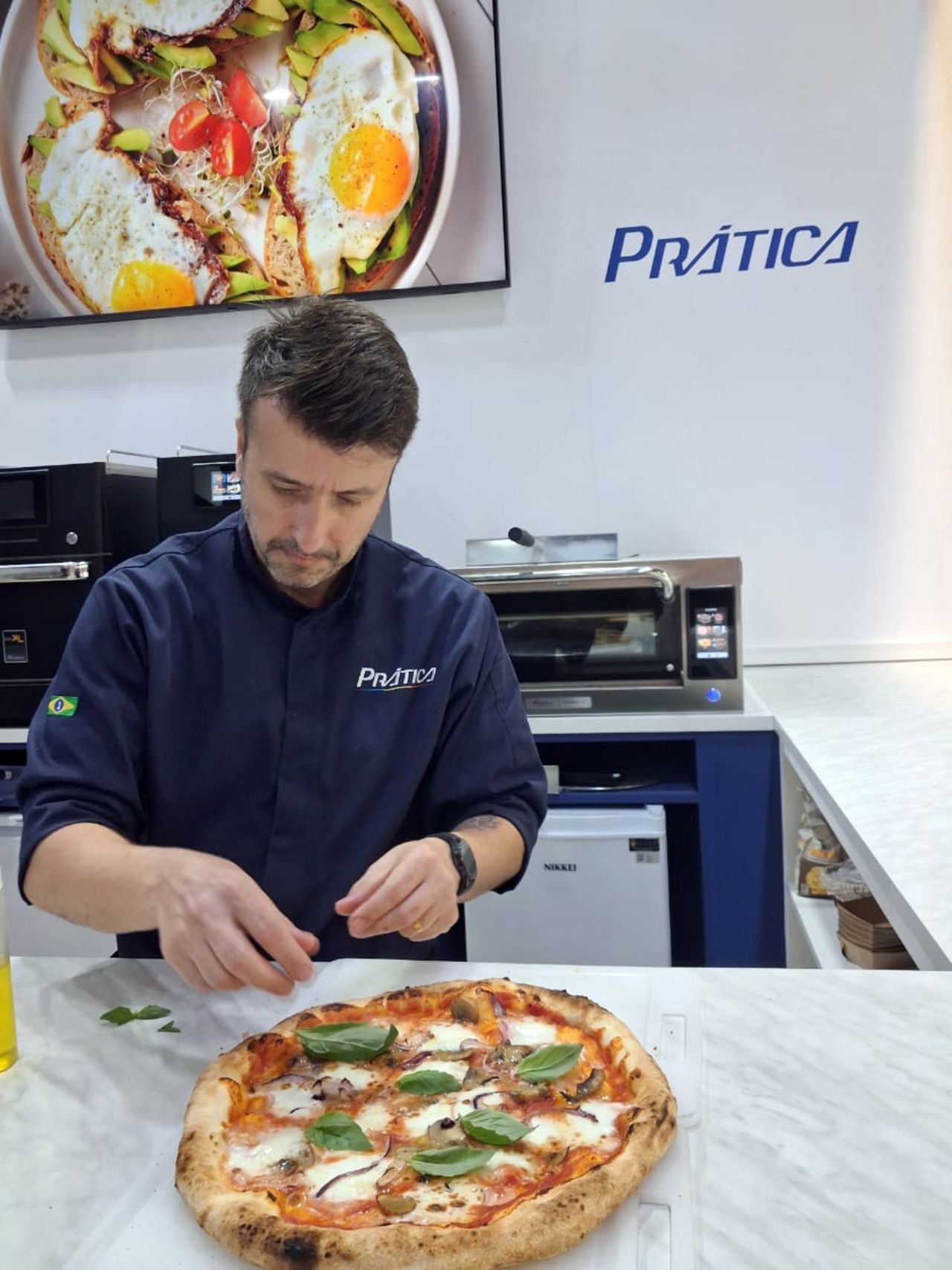 Chef garnishes a pizza with basil. A display shows a cooked dish. 