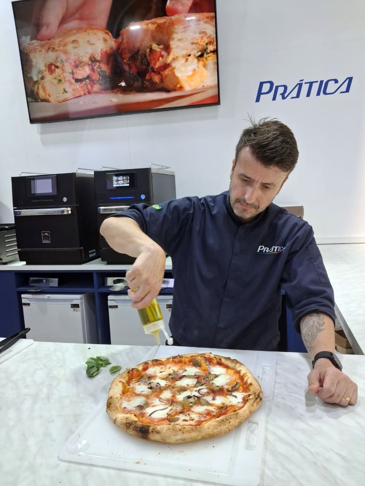 Chef adding oil to pizza, in front of ovens. Sign reads 