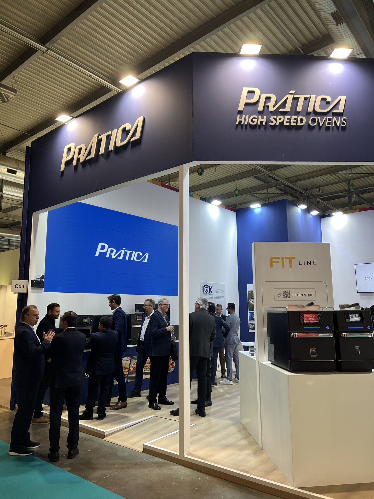 Booth for Prática High Speed Ovens at a trade show; several people in suits stand around the display.