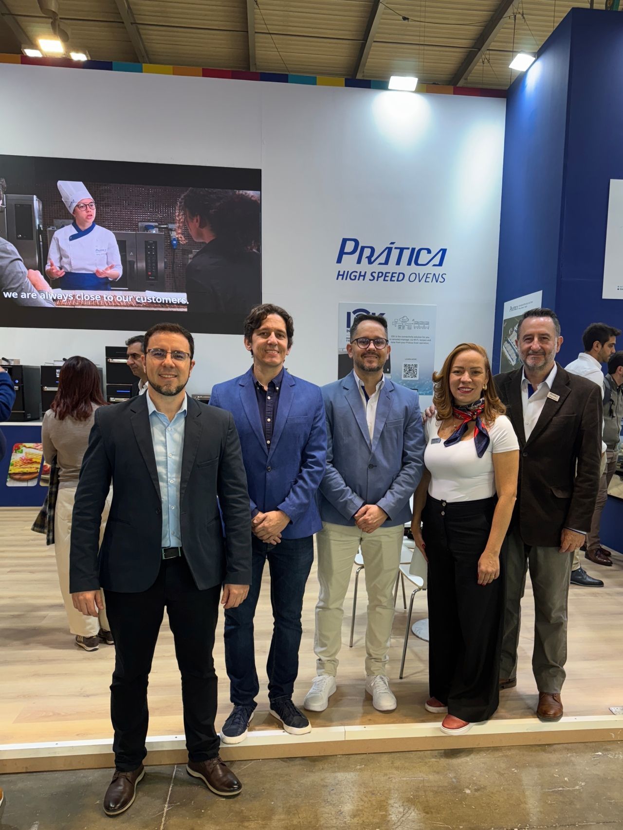 Five people posing in front of a booth with a logo and screen; indoors, neutral colors.