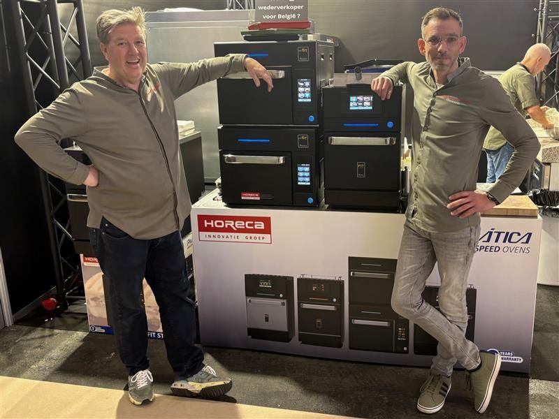 Two men pose with commercial ovens at a trade show booth. One man leans on the ovens, other on a sign.