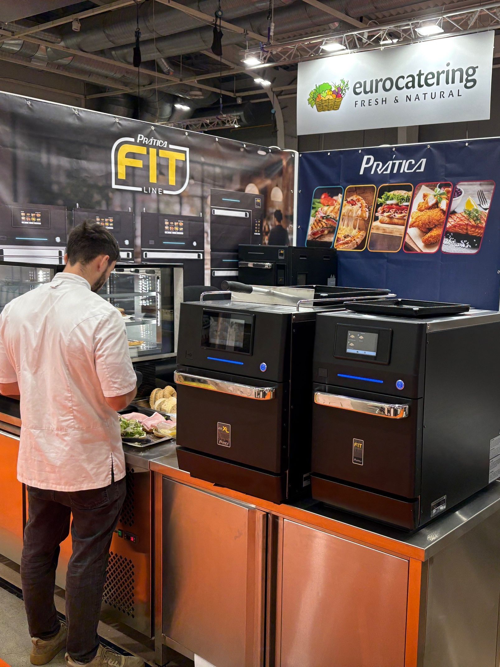 Man preparing food at a catering expo booth with two black commercial kitchen appliances.