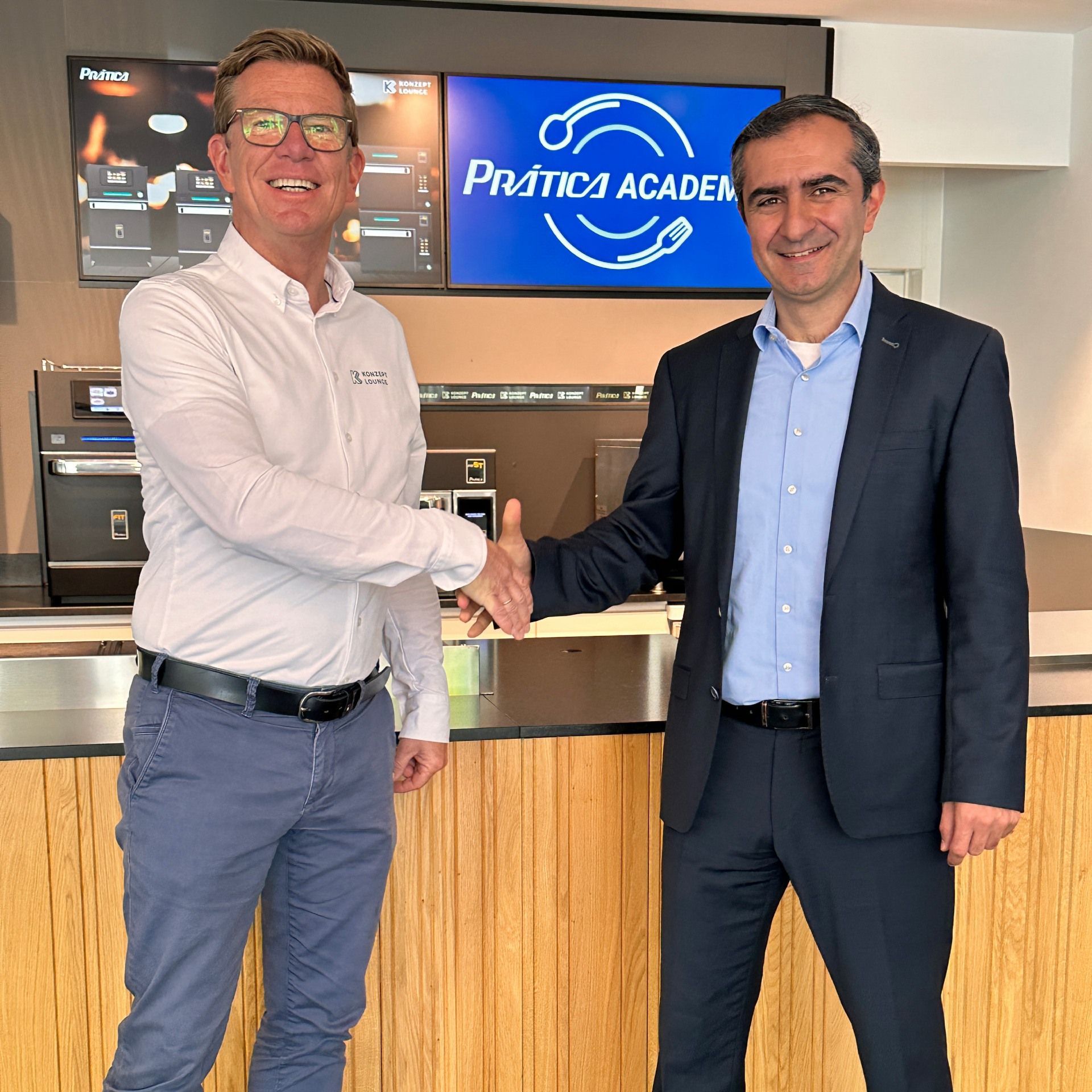 Two men shaking hands in front of a sign that says pratico academ