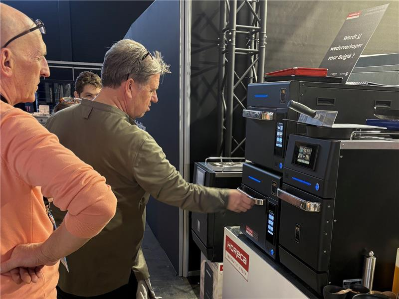 Two men looking at black electronic equipment at a trade show booth. One man is operating a machine.