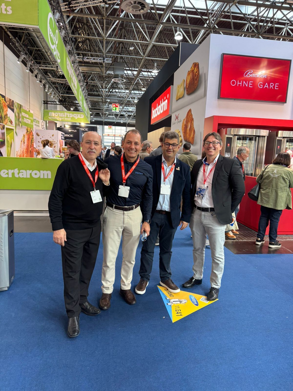Four men posing at a trade show booth. Smiling, wearing lanyards. Show setting, booths & people in background.
