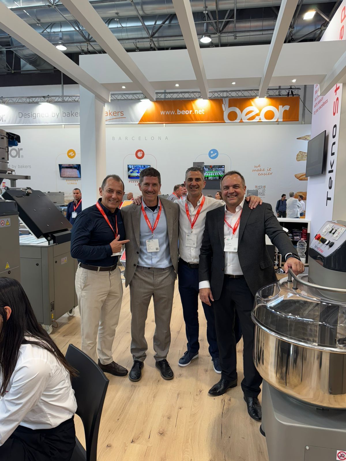 Four men smiling at a trade show booth, with baking equipment and the