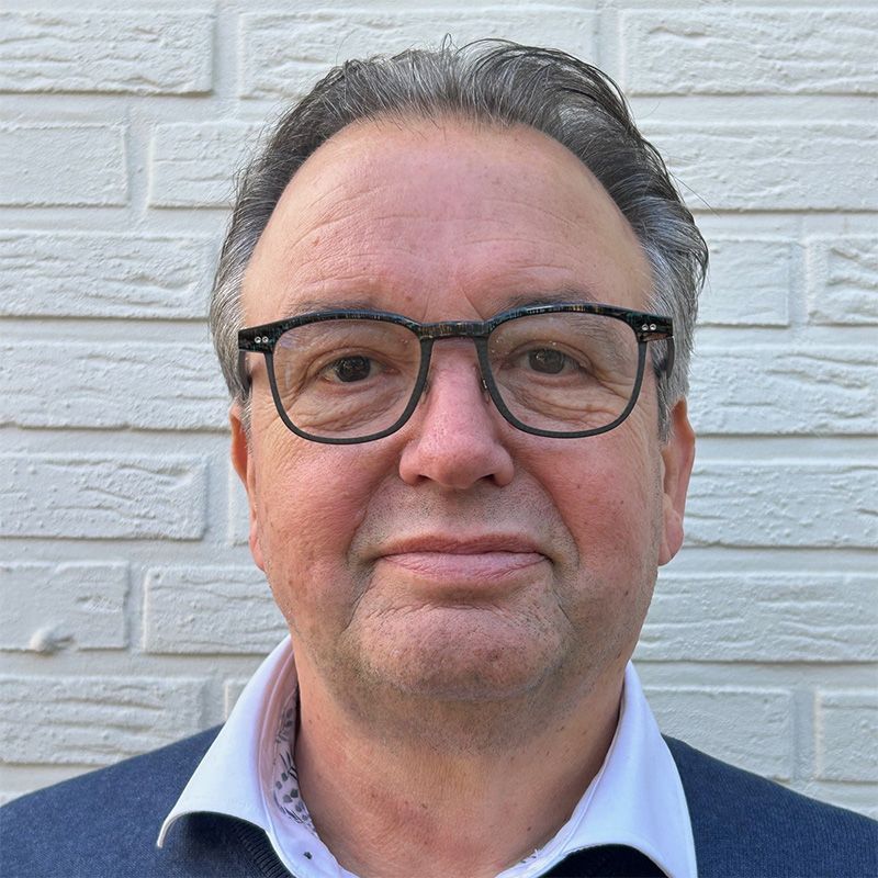 A man wearing glasses and a blue shirt is standing in front of a white brick wall.