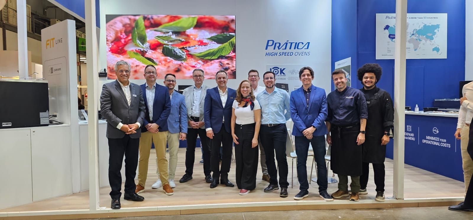 A group of people posing in front of a booth at a trade show. A large screen displays food.