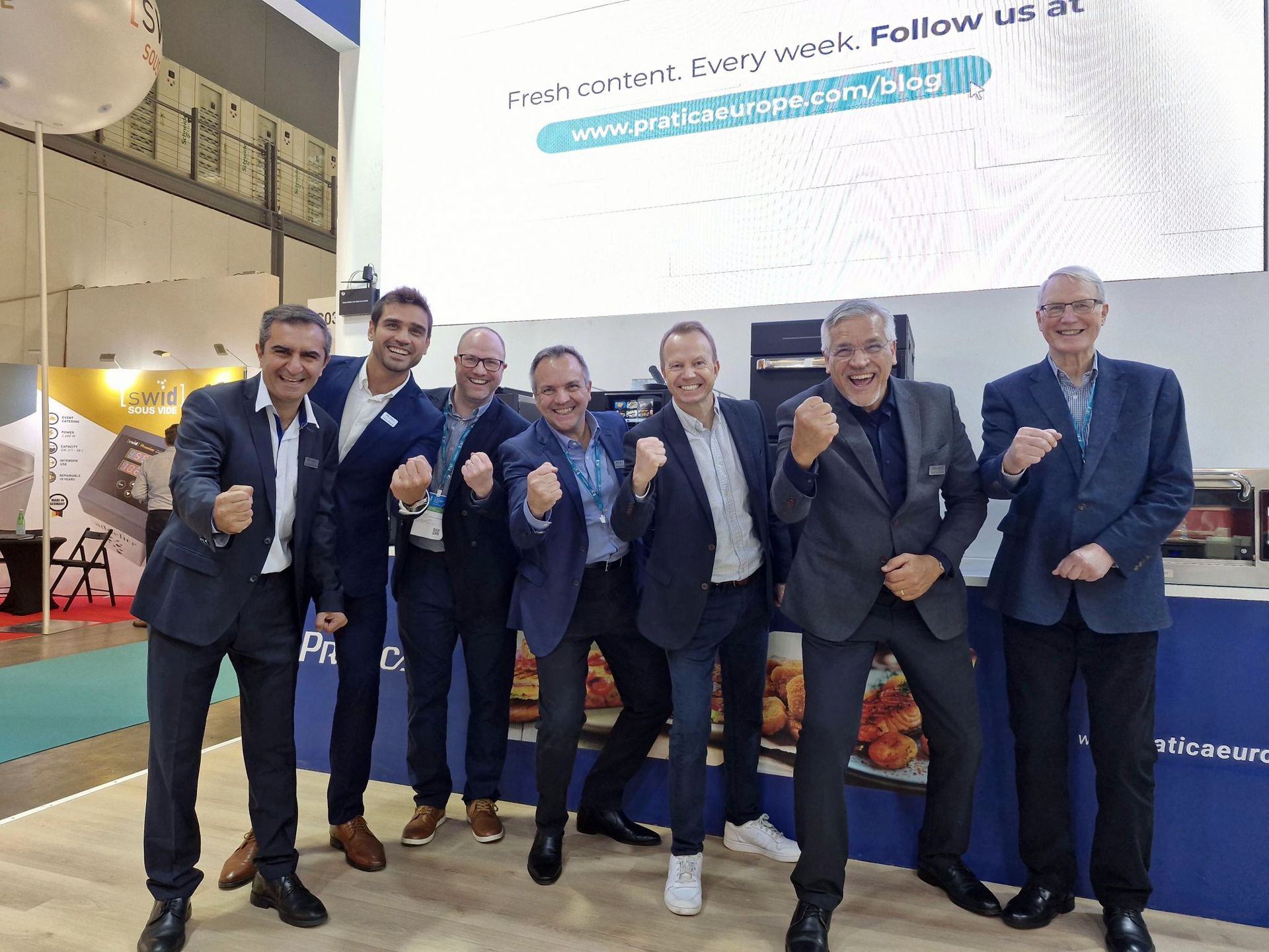 Seven men in suits cheering in front of a display at an event.