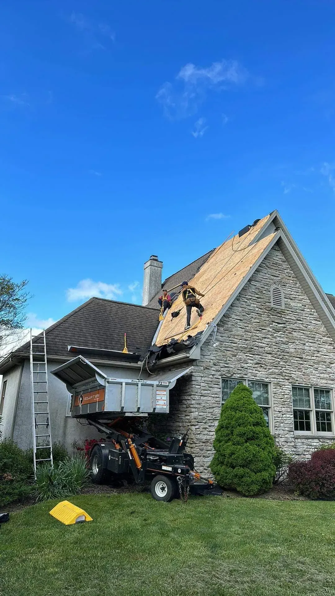 Roofers on a house with stone siding replacing shingles. A lift and ladder are present on the lawn.