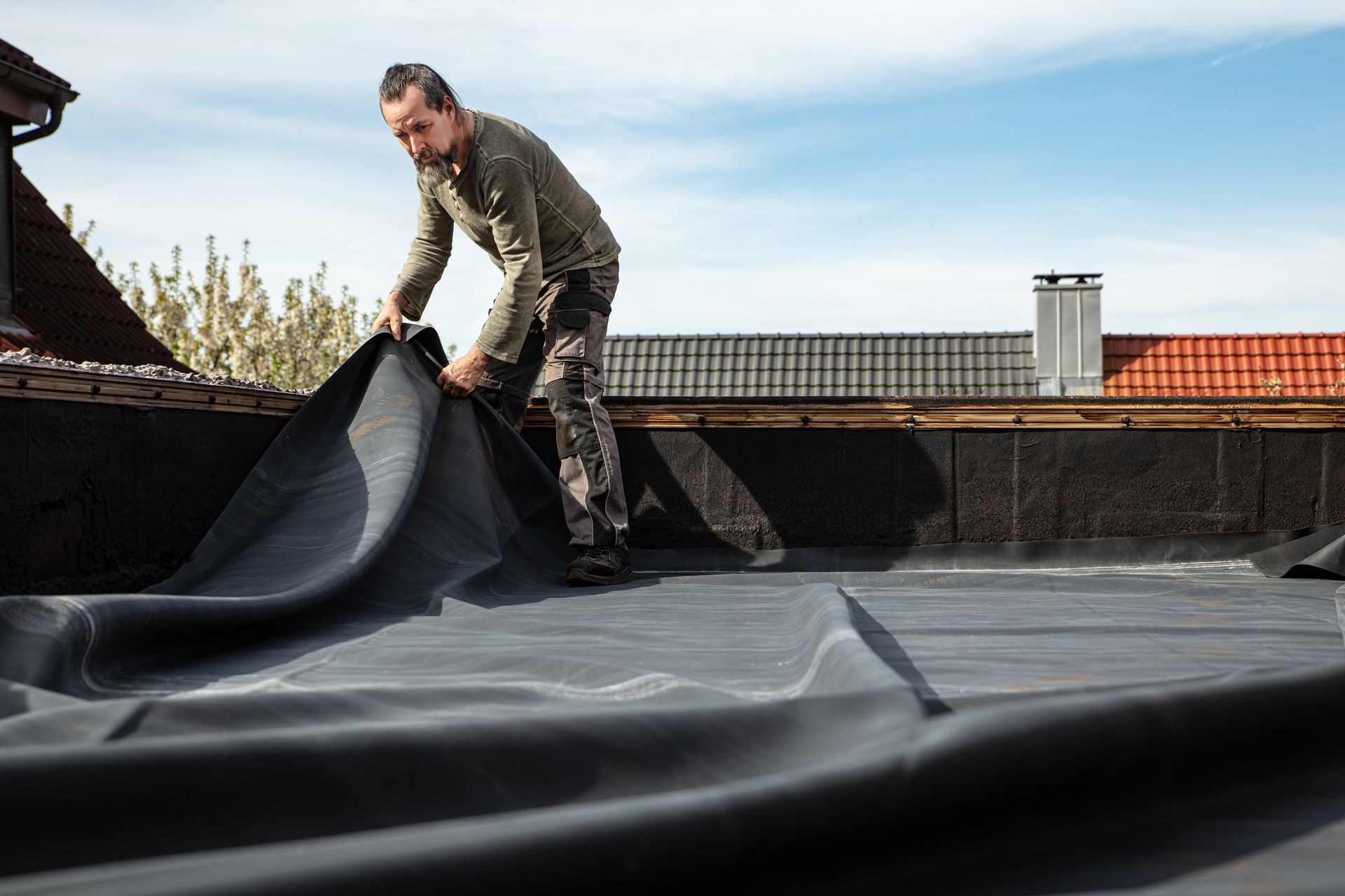 Man on a rooftop installing black roofing material under a blue sky.