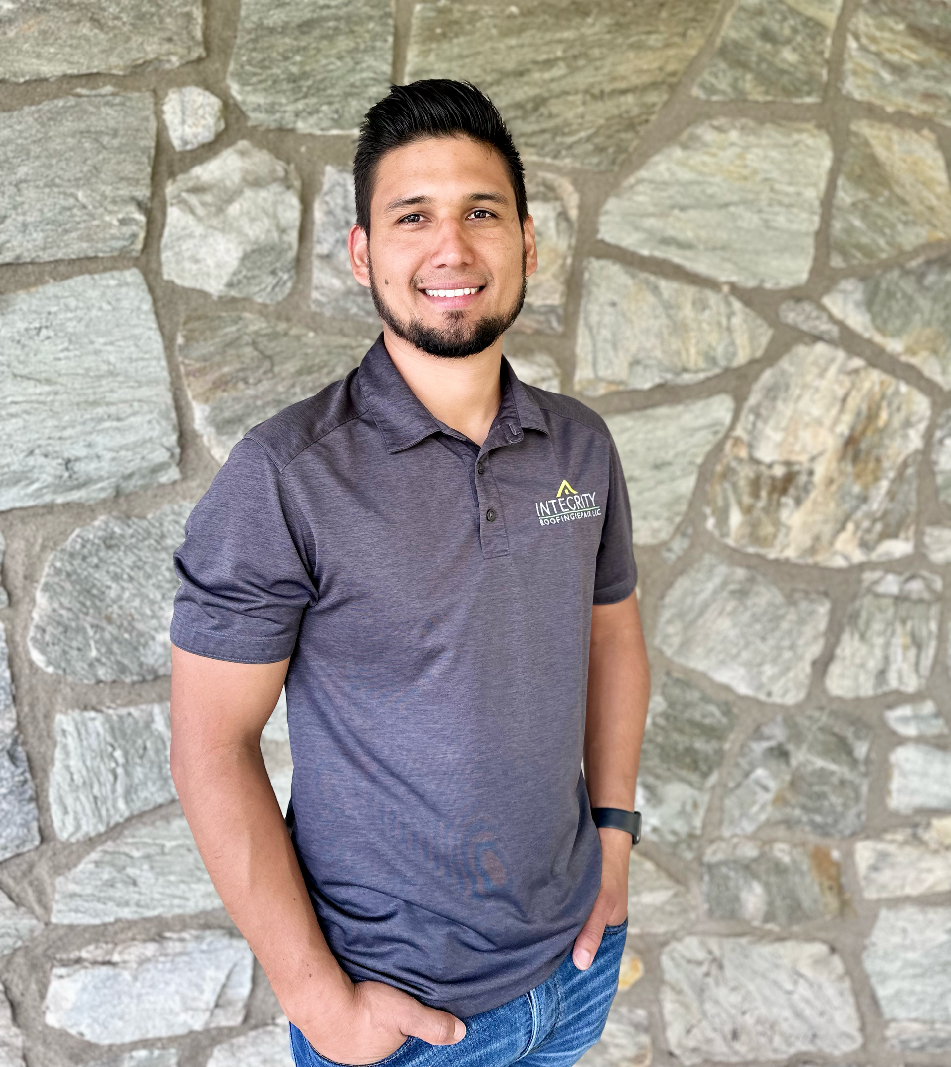 Man in gray polo shirt smiling, standing in front of a stone wall, hands in pockets.