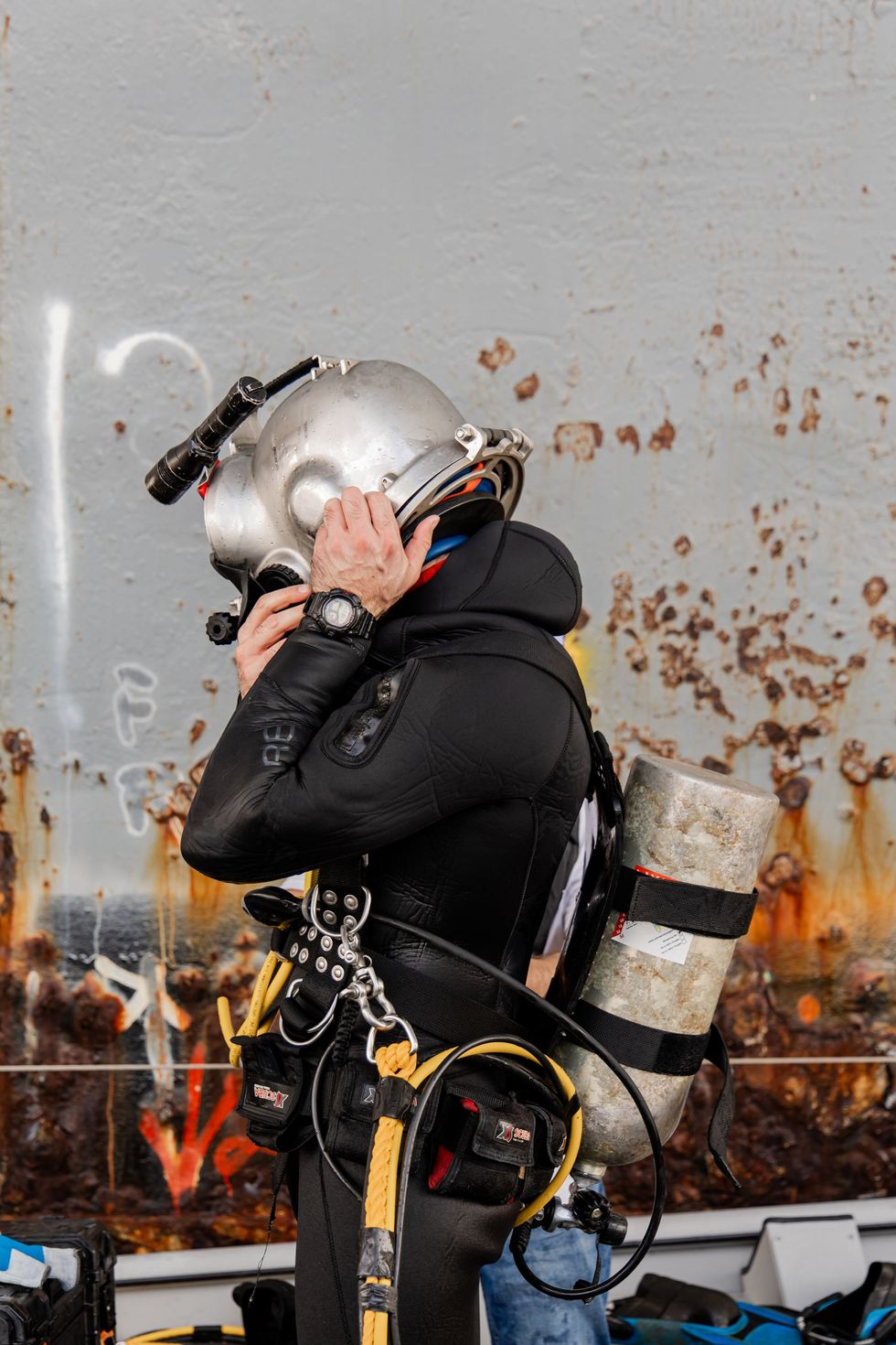 Diver in silver helmet and black wetsuit adjusts face mask in front of a ship