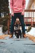 Person standing on a porch with a small black dog between their legs, snow on the ground.