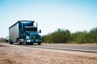 Blue semi-truck driving on a road with green foliage and a clear blue sky.