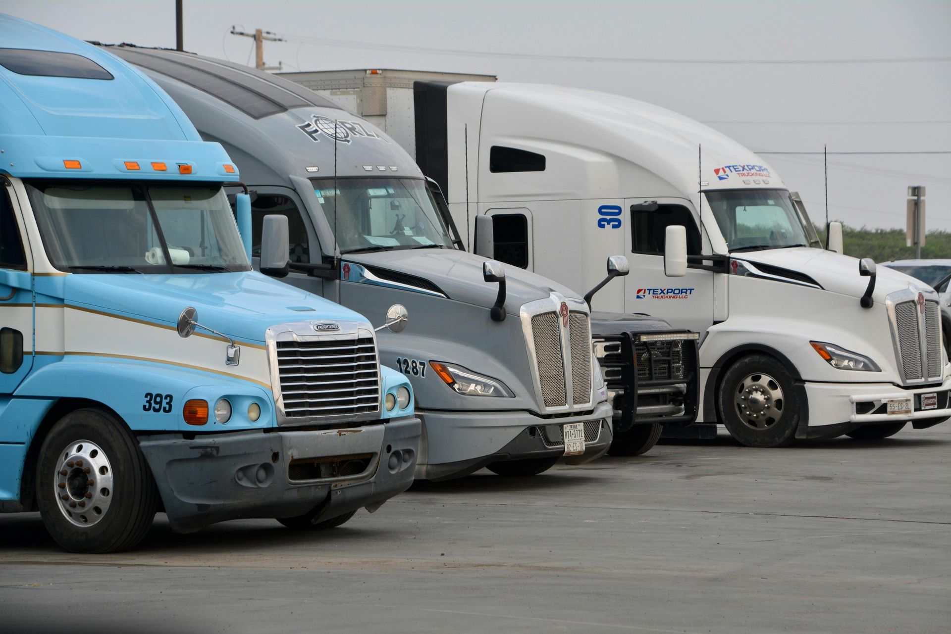 Three semi-trucks parked in a lot; light blue, gray, and white.