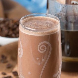 Chocolate-colored drink in a clear glass with swirls, coffee beans and a carafe in the background.