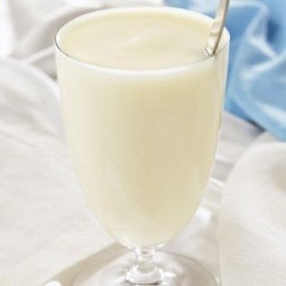 Glass of thick, creamy white beverage with a spoon in it, on a white surface.