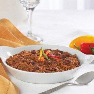 Chili in a white bowl with peppers on a white table, next to a spoon, cloth and glass.