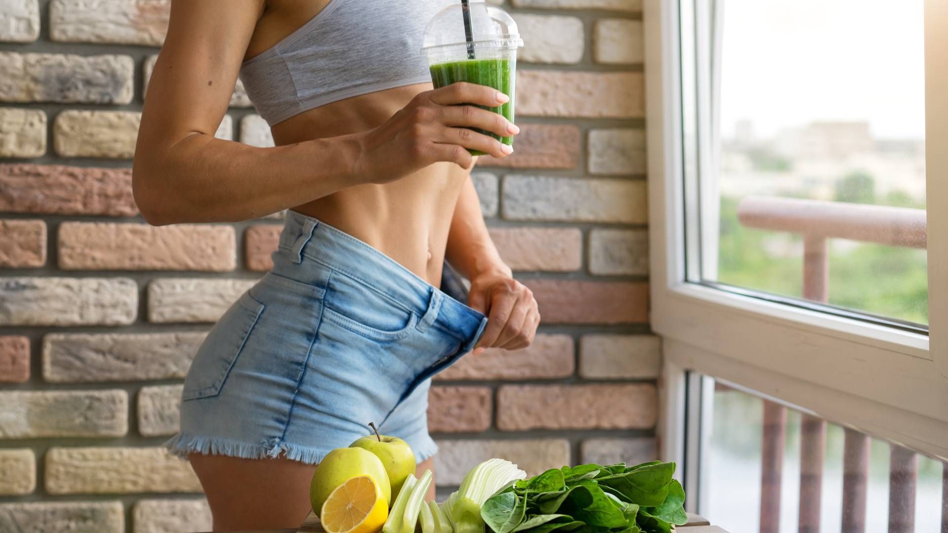 Woman in shorts, showing weight loss, holding smoothie near window.