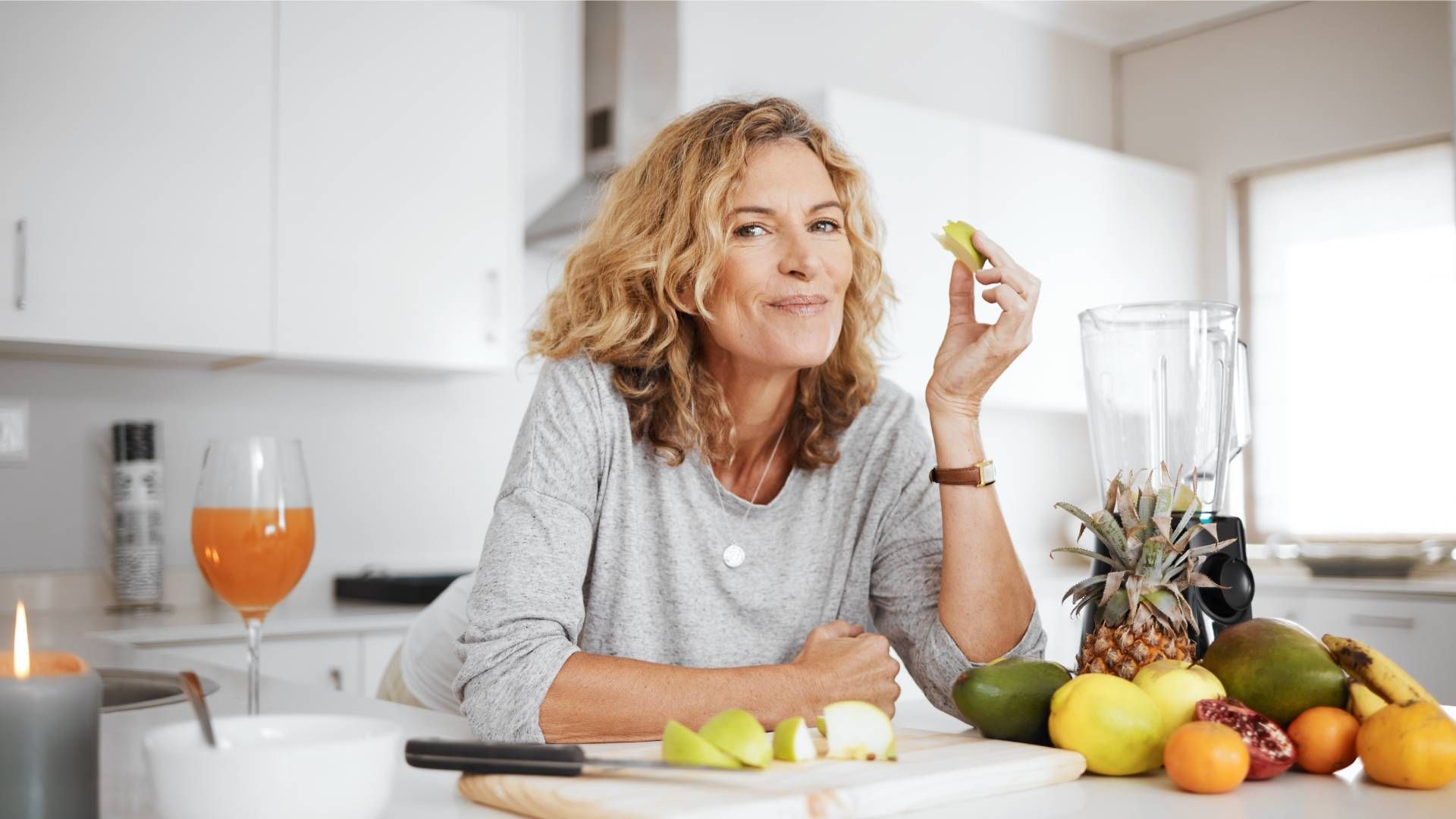 Woman in kitchen holding lime, surrounded by fruit, smiling.