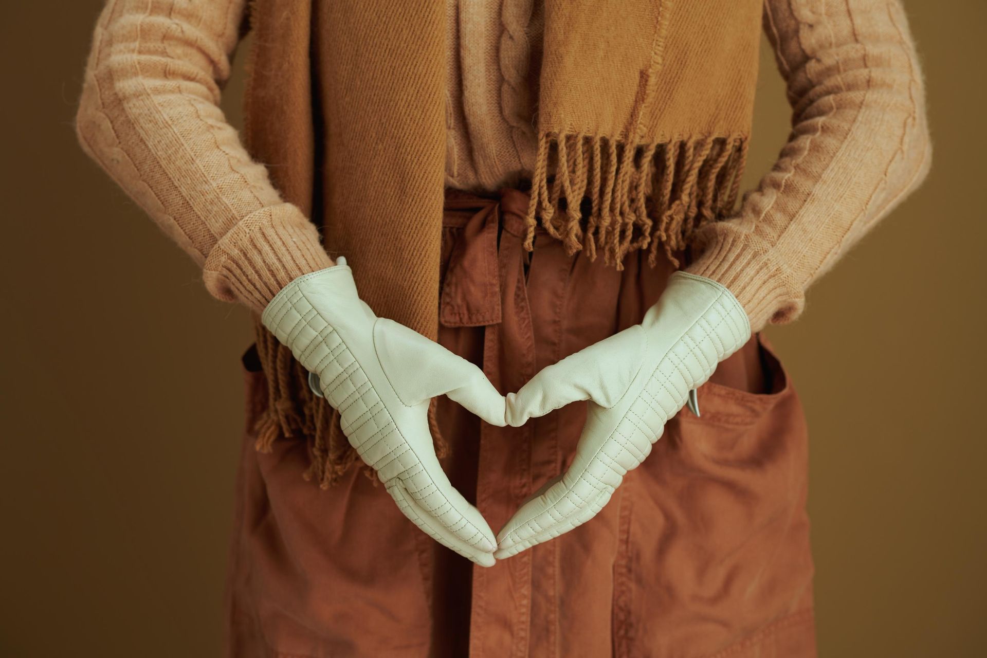 Person in a beige sweater and brown pants forming a heart shape with mint green gloved hands
