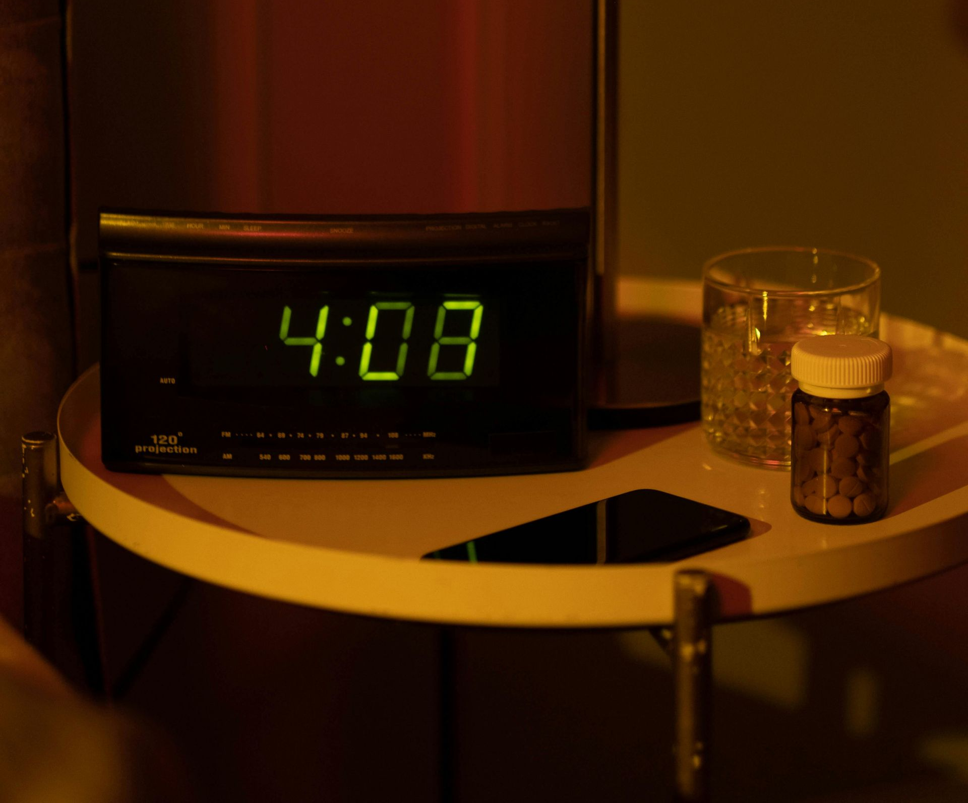 Clock showing 4:08 on a nightstand with a glass of water, pills, and a phone.