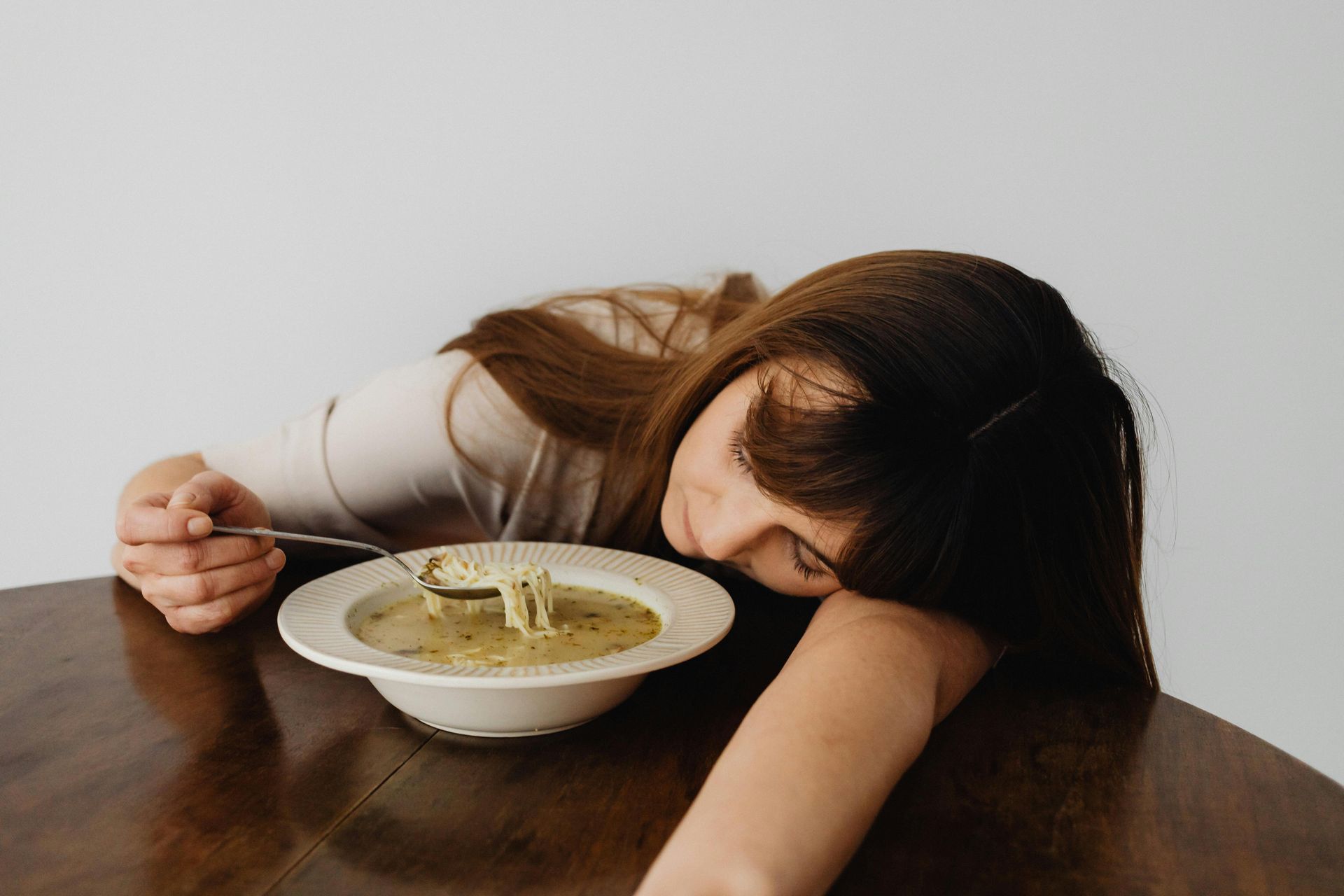 Woman slumped on a table, head next to a bowl of soup, spoon in hand, appearing tired.